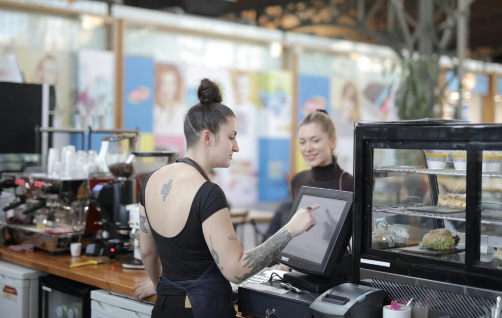 Female cashier with tattoos helps a customer with a transaction at a cafe counter.